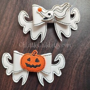 May include: Two white fabric hair bows with embroidered designs. One bow features a white ghost with a black face and orange nose. The other bow features an orange pumpkin with a black face.