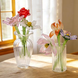 Peut inclure: Deux vases en verre transparent avec des fleurs en verre color&eacute;es. L'un contient une rose rouge, une tulipe rose et une fleur blanche. L'autre pr&eacute;sente un lis orange, une tulipe rose et une fleur violette. Les vases sont sur une table marron clair.