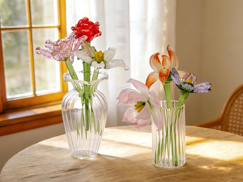 Peut inclure: Deux vases en verre transparent avec des fleurs en verre color&eacute;es. L'un contient une rose rouge, une tulipe rose et une fleur blanche. L'autre pr&eacute;sente un lis orange, une tulipe rose et une fleur violette. Les vases sont sur une table marron clair.