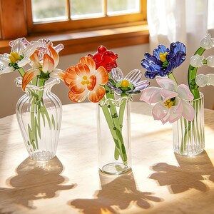Peut inclure: Trois vases en verre avec des fleurs en verre color&eacute;es. Les fleurs comprennent des fleurs orange, rouges, bleues et roses. Les vases sont transparents et pos&eacute;s sur une table de couleur claire. Les fleurs sont dispos&eacute;es en bouquet.