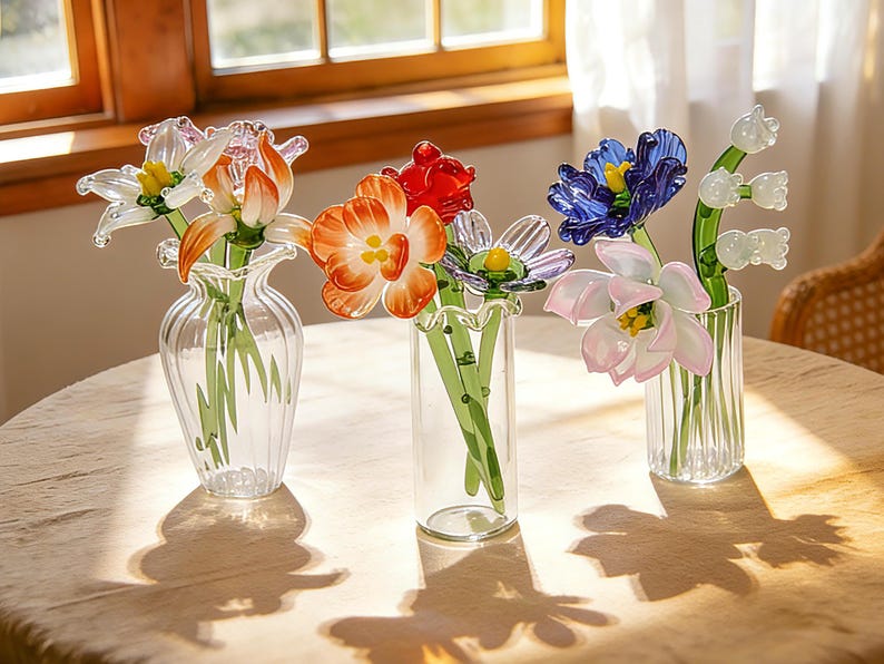 Peut inclure: Trois vases en verre avec des fleurs en verre color&eacute;es. Les fleurs comprennent des fleurs orange, rouges, bleues et roses. Les vases sont transparents et pos&eacute;s sur une table de couleur claire. Les fleurs sont dispos&eacute;es en bouquet.