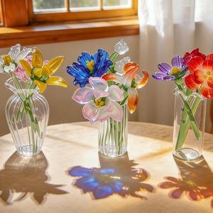 Peut inclure: Trois arrangements floraux en verre dans des vases transparents sont pos&eacute;s sur une table ronde. Les fleurs sont de couleurs vari&eacute;es, dont le jaune, le bleu, l'orange, le rouge et le violet. Les vases sont transparents et ont un dessus &agrave; volants. La table est de couleur claire, et l'arri&egrave;re-plan comprend une fen&ecirc;tre et une chaise.