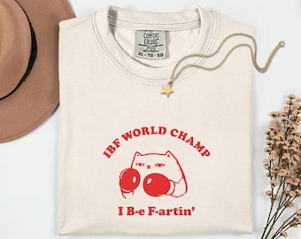 May include: Cream-colored t-shirt with red graphic text and a cat wearing boxing gloves. The text reads "IBF WORLD CHAMP" above the cat and "I B-e F-artin'" below. A gold star pendant necklace and a brown hat are also in the image.