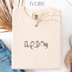 Puede incluir: Camiseta de color marfil con cuello redondo y mangas cortas. La camiseta presenta un gr&aacute;fico de cuatro nutrias de dibujos animados en varias poses. Un collar dorado con un peque&ntilde;o colgante est&aacute; drapeado sobre la camiseta. La camiseta est&aacute; doblada sobre una superficie blanca.