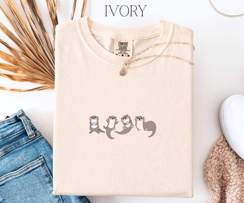 Puede incluir: Camiseta de color marfil con cuello redondo y mangas cortas. La camiseta presenta un gr&aacute;fico de cuatro nutrias de dibujos animados en varias poses. Un collar dorado con un peque&ntilde;o colgante est&aacute; drapeado sobre la camiseta. La camiseta est&aacute; doblada sobre una superficie blanca.