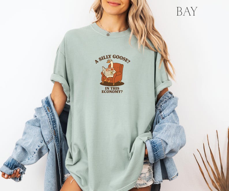 Puede incluir: Camiseta verde claro con el texto "A SILLY GOOSE? IN THIS ECONOMY?" y un dibujo de un ganso leyendo un peri&oacute;dico en un sill&oacute;n marr&oacute;n. La camiseta tiene mangas cortas y un corte holgado.