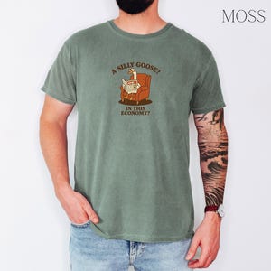 Puede incluir: Camiseta verde musgo con un gr&aacute;fico de un ganso leyendo un peri&oacute;dico en una silla. El texto de la camiseta dice "A SILLY GOOSE? IN THIS ECONOMY?" La camiseta est&aacute; hecha de un material suave y c&oacute;modo.