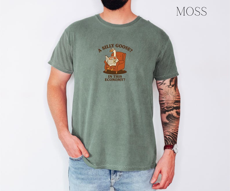 Puede incluir: Camiseta verde musgo con un gr&aacute;fico de un ganso leyendo un peri&oacute;dico en una silla. El texto de la camiseta dice "A SILLY GOOSE? IN THIS ECONOMY?" La camiseta est&aacute; hecha de un material suave y c&oacute;modo.