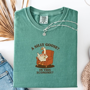 Puede incluir: Camiseta verde azulado con la frase "A silly goose? In this economy?" y un dibujo de un ganso leyendo un peri&oacute;dico. Un collar plateado con un colgante est&aacute; sobre la camiseta. La camiseta est&aacute; doblada sobre una superficie blanca.