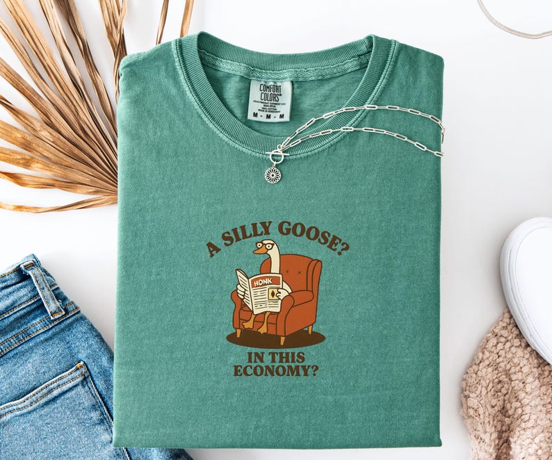 Puede incluir: Camiseta verde azulado con la frase "A silly goose? In this economy?" y un dibujo de un ganso leyendo un peri&oacute;dico. Un collar plateado con un colgante est&aacute; sobre la camiseta. La camiseta est&aacute; doblada sobre una superficie blanca.