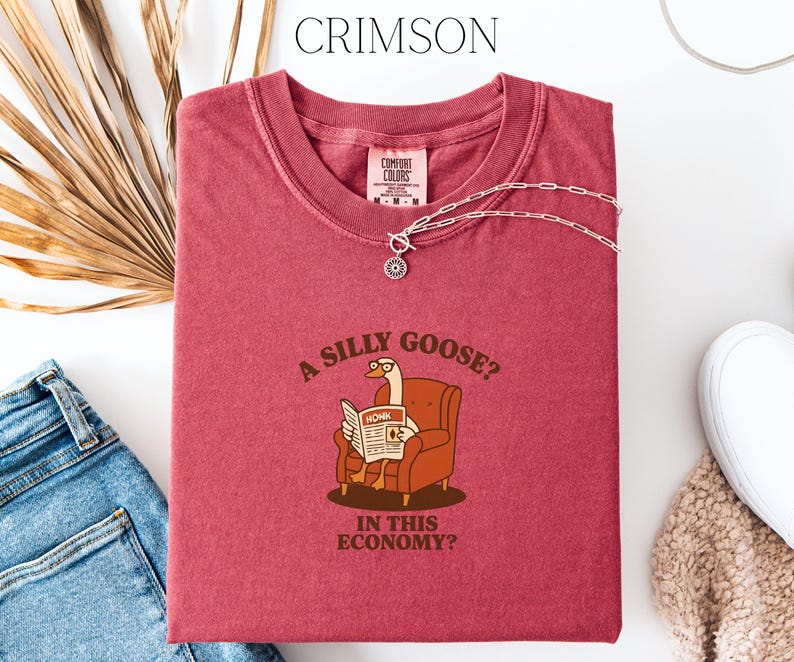 Puede incluir: Camiseta de color carmes&iacute; con el texto "A silly goose? In this economy?" y un dibujo de un ganso leyendo un peri&oacute;dico. Un collar plateado con un colgante circular est&aacute; sobre la camiseta. La camiseta est&aacute; doblada sobre una superficie blanca.