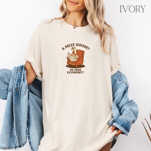Puede incluir: Camiseta de color marfil con un dibujo de un ganso leyendo un peri&oacute;dico en un sill&oacute;n marr&oacute;n. El texto de la camiseta dice: "A SILLY GOOSE? IN THIS ECONOMY?" La camiseta tiene un corte holgado.