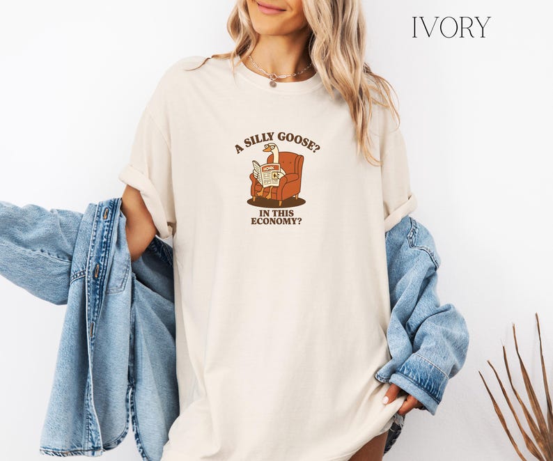 Puede incluir: Camiseta de color marfil con un dibujo de un ganso leyendo un peri&oacute;dico en un sill&oacute;n marr&oacute;n. El texto de la camiseta dice: "A SILLY GOOSE? IN THIS ECONOMY?" La camiseta tiene un corte holgado.