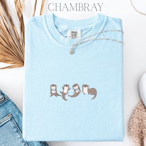 Puede incluir: Una camiseta azul claro con la palabra "CHAMBRAY" en la parte superior. La camiseta presenta un dise&ntilde;o bordado de cuatro nutrias. Un collar plateado con un peque&ntilde;o colgante est&aacute; sobre la camiseta.