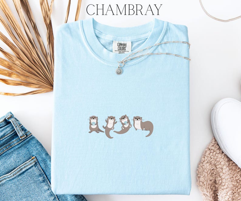 Puede incluir: Una camiseta azul claro con la palabra "CHAMBRAY" en la parte superior. La camiseta presenta un dise&ntilde;o bordado de cuatro nutrias. Un collar plateado con un peque&ntilde;o colgante est&aacute; sobre la camiseta.