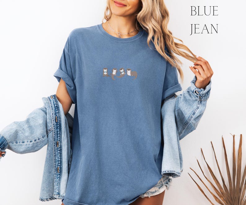 Puede incluir: Camiseta azul vaquero con cuello redondo y mangas cortas. La camiseta presenta un dise&ntilde;o bordado de cuatro nutrias. Las palabras "BLUE JEAN" est&aacute;n impresas en una fuente serif en la esquina superior derecha.