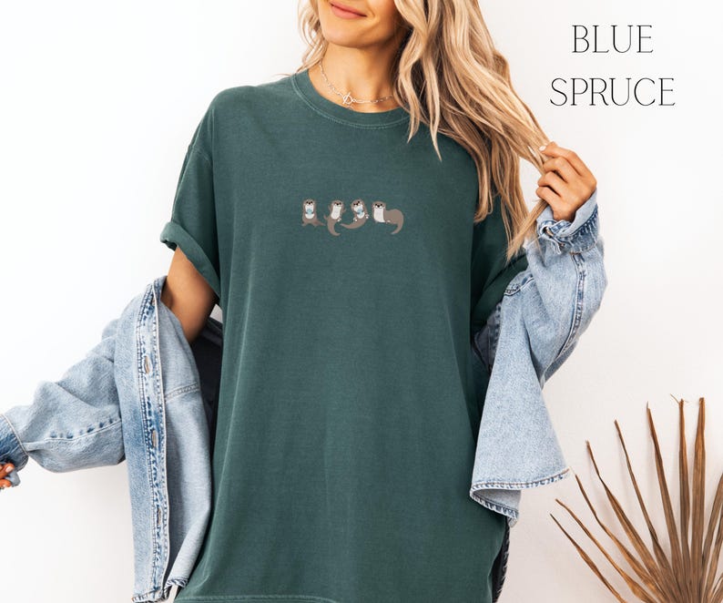 Puede incluir: Una camiseta verde oscuro con un peque&ntilde;o dise&ntilde;o bordado de nutrias. La camiseta tiene las mangas enrolladas y se combina con una chaqueta vaquera. Las palabras "BLUE SPRUCE" est&aacute;n impresas en negro en la parte superior derecha.