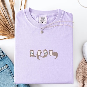 Puede incluir: Una camiseta lila Comfort Colors con un dise&ntilde;o de cuatro nutrias. Cada nutria del gr&aacute;fico sostiene un peque&ntilde;o objeto azul. Un collar plateado con un colgante de sol est&aacute; sobre la camiseta. La camiseta est&aacute; doblada sobre una superficie blanca.