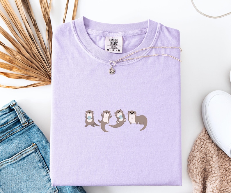 Puede incluir: Una camiseta lila Comfort Colors con un dise&ntilde;o de cuatro nutrias. Cada nutria del gr&aacute;fico sostiene un peque&ntilde;o objeto azul. Un collar plateado con un colgante de sol est&aacute; sobre la camiseta. La camiseta est&aacute; doblada sobre una superficie blanca.
