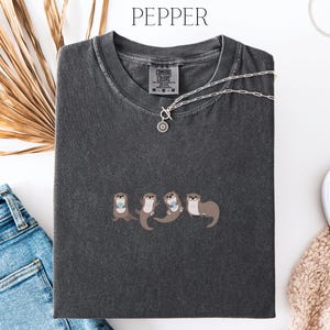 Puede incluir: Una camiseta gris oscuro con un dise&ntilde;o bordado de cuatro nutrias tomadas de la mano. La camiseta tiene cuello redondo y mangas cortas. Un collar plateado con un colgante circular descansa sobre la camiseta. La palabra "PEPPER" est&aacute; arriba.