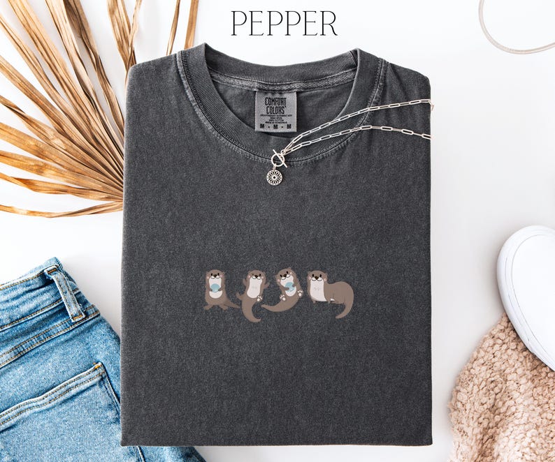 Puede incluir: Una camiseta gris oscuro con un dise&ntilde;o bordado de cuatro nutrias tomadas de la mano. La camiseta tiene cuello redondo y mangas cortas. Un collar plateado con un colgante circular descansa sobre la camiseta. La palabra "PEPPER" est&aacute; arriba.