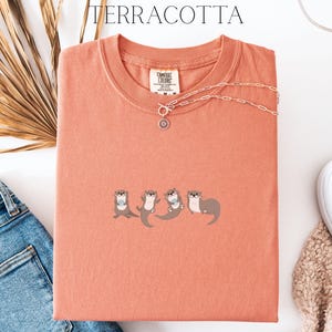 Puede incluir: Camiseta de color terracota con cuello redondo y mangas cortas. La camiseta presenta un dise&ntilde;o bordado de cuatro nutrias. Un collar plateado con un colgante est&aacute; drapeado sobre la camiseta. La palabra "TERRACOTTA" est&aacute; impresa en la parte superior.