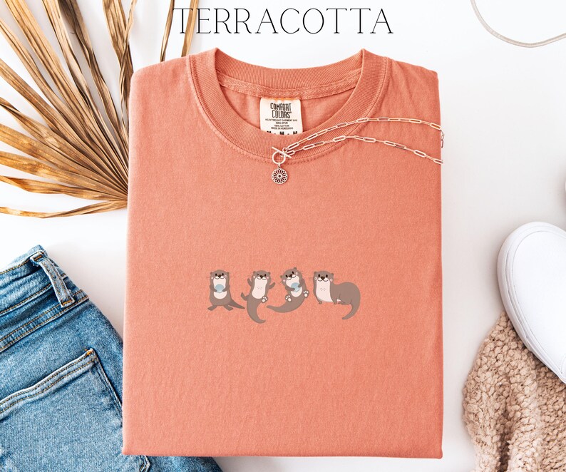 Puede incluir: Camiseta de color terracota con cuello redondo y mangas cortas. La camiseta presenta un dise&ntilde;o bordado de cuatro nutrias. Un collar plateado con un colgante est&aacute; drapeado sobre la camiseta. La palabra "TERRACOTTA" est&aacute; impresa en la parte superior.