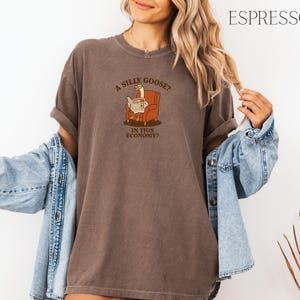 Puede incluir: Camiseta color espresso con la frase "A silly goose? In this economy?" impresa en la parte delantera. El gr&aacute;fico presenta un ganso leyendo un peri&oacute;dico en un sill&oacute;n marr&oacute;n. La camiseta tiene mangas cortas y un corte holgado.