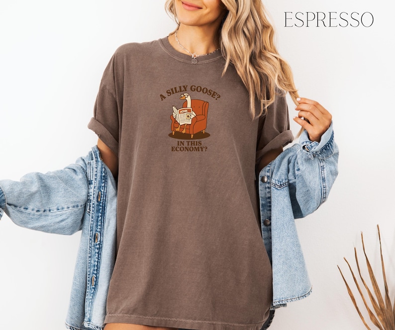 Puede incluir: Camiseta color espresso con la frase "A silly goose? In this economy?" impresa en la parte delantera. El gr&aacute;fico presenta un ganso leyendo un peri&oacute;dico en un sill&oacute;n marr&oacute;n. La camiseta tiene mangas cortas y un corte holgado.