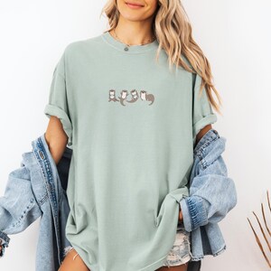 Puede incluir: Una camiseta verde claro con un dise&ntilde;o bordado de nutrias. Las nutrias est&aacute;n representadas en fila, con los brazos entrelazados. La camiseta tiene un corte holgado y mangas cortas.