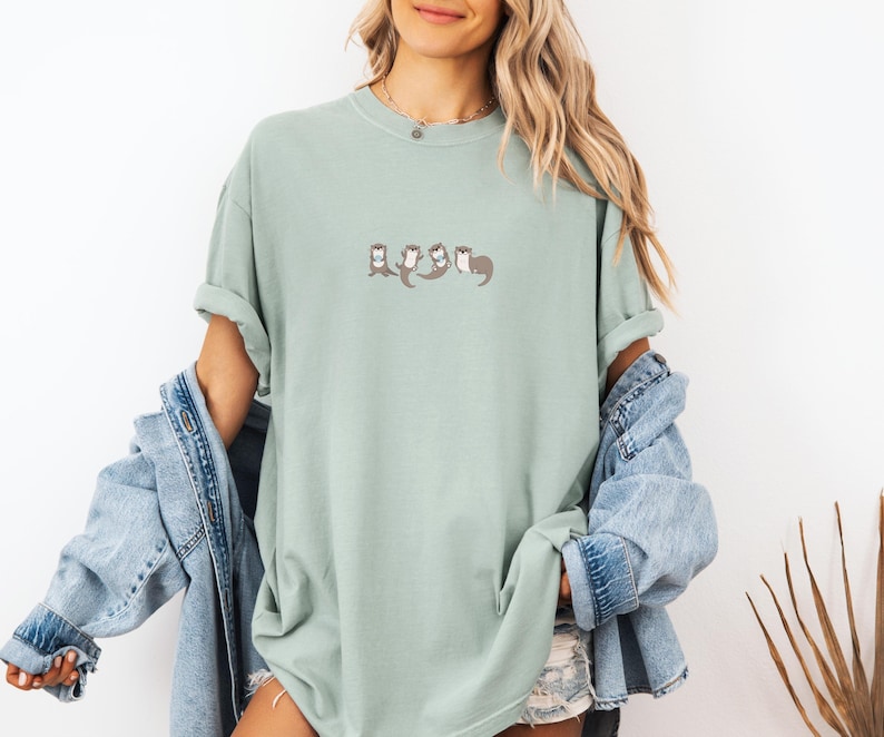 Puede incluir: Una camiseta verde claro con un dise&ntilde;o bordado de nutrias. Las nutrias est&aacute;n representadas en fila, con los brazos entrelazados. La camiseta tiene un corte holgado y mangas cortas.