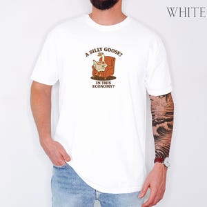 Puede incluir: Camiseta blanca con un dibujo de un ganso leyendo un peri&oacute;dico en un sill&oacute;n marr&oacute;n. El texto de la camiseta dice: "A silly goose? In this economy?" La palabra "WHITE" est&aacute; impresa en la esquina superior derecha.
