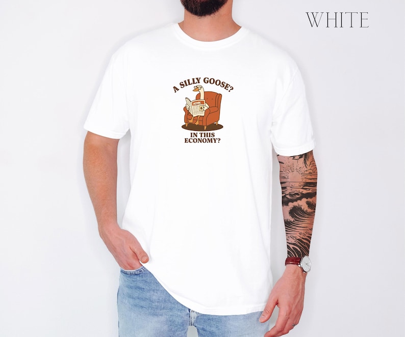 Puede incluir: Camiseta blanca con un dibujo de un ganso leyendo un peri&oacute;dico en un sill&oacute;n marr&oacute;n. El texto de la camiseta dice: "A silly goose? In this economy?" La palabra "WHITE" est&aacute; impresa en la esquina superior derecha.