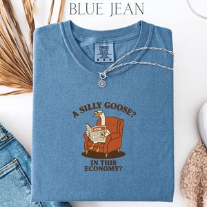 Puede incluir: Camiseta azul Comfort Colors con el texto "A SILLY GOOSE? IN THIS ECONOMY?" y un dibujo de un ganso leyendo un peri&oacute;dico. Un collar plateado est&aacute; sobre la camiseta. La camiseta se combina con vaqueros azules y un su&eacute;ter beige.