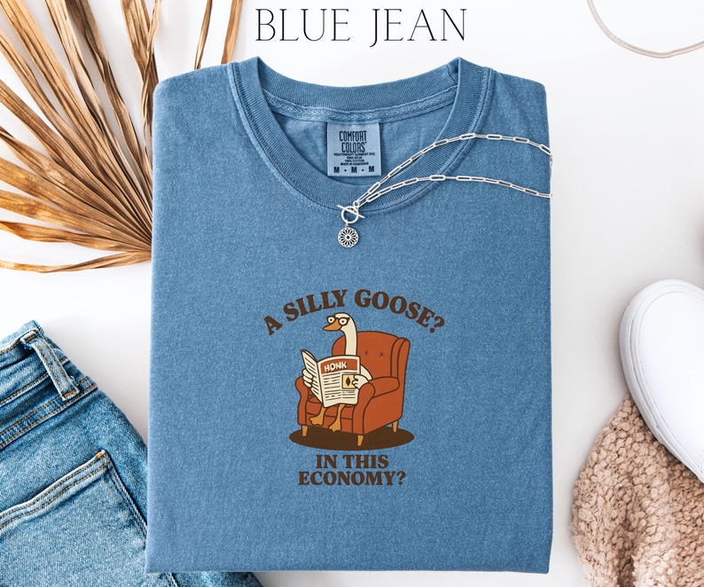 Puede incluir: Camiseta azul Comfort Colors con el texto "A SILLY GOOSE? IN THIS ECONOMY?" y un dibujo de un ganso leyendo un peri&oacute;dico. Un collar plateado est&aacute; sobre la camiseta. La camiseta se combina con vaqueros azules y un su&eacute;ter beige.