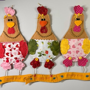 May include: Three handmade chicken-shaped kitchen organizers. Each has a tan body, a yellow beak, and a red wattle. They wear patterned aprons and have a yellow banner that reads "Grocery List".