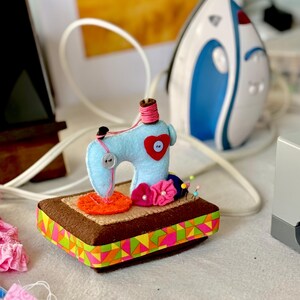 Op de afbeelding: Een lichtblauwe vilten naaimachine met een rood hart en een roze garenklos staat op een bruine rechthoekige basis met een kleurrijke geometrische lintafwerking. Een wit en blauw strijkijzer staat op de achtergrond.