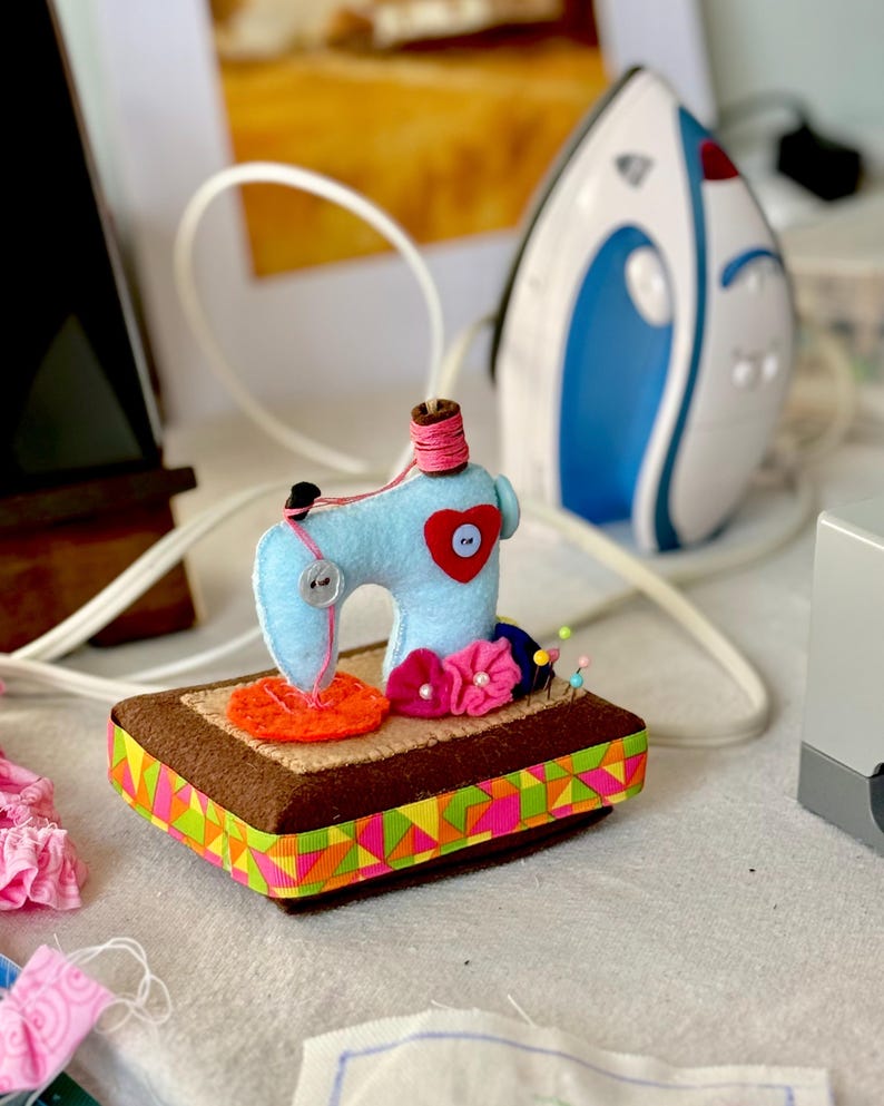 Op de afbeelding: Een lichtblauwe vilten naaimachine met een rood hart en een roze garenklos staat op een bruine rechthoekige basis met een kleurrijke geometrische lintafwerking. Een wit en blauw strijkijzer staat op de achtergrond.