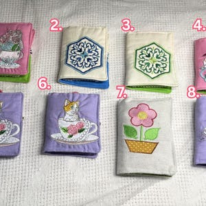 May include: A collection of eight small, fabric-covered books with embroidered designs. The books feature various motifs, including flowers, cats in teacups, and geometric patterns. Some books have the word "sewing" embroidered on them.