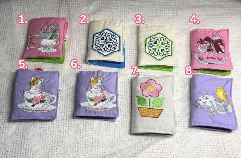 May include: A collection of eight small, fabric-covered books with embroidered designs. The books feature various motifs, including flowers, cats in teacups, and geometric patterns. Some books have the word "sewing" embroidered on them.
