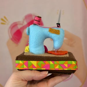 Op de afbeelding: Een handgemaakte miniatuur naaimachine van blauw vilt, met een roze garenklos en oranje stoffen details. De basis is bruin met een kleurrijke geometrische lintafwerking. Een charmant decoratief item.