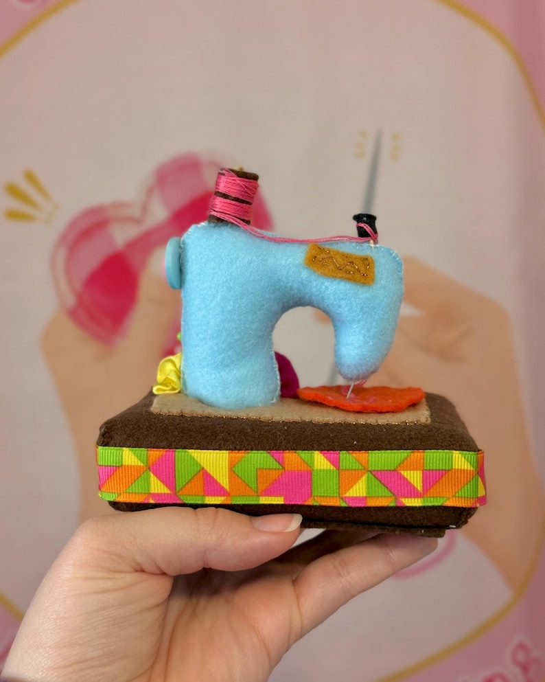 Op de afbeelding: Een handgemaakte miniatuur naaimachine van blauw vilt, met een roze garenklos en oranje stoffen details. De basis is bruin met een kleurrijke geometrische lintafwerking. Een charmant decoratief item.