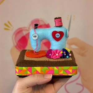Op de afbeelding: Een handgemaakte vilten naaimachine in lichtblauw, met een rood hartdetail en een spoel roze garen. Het staat op een bruine basis met een kleurrijke geometrische lintafwerking en een klein speldenkussen met bloemspelden.