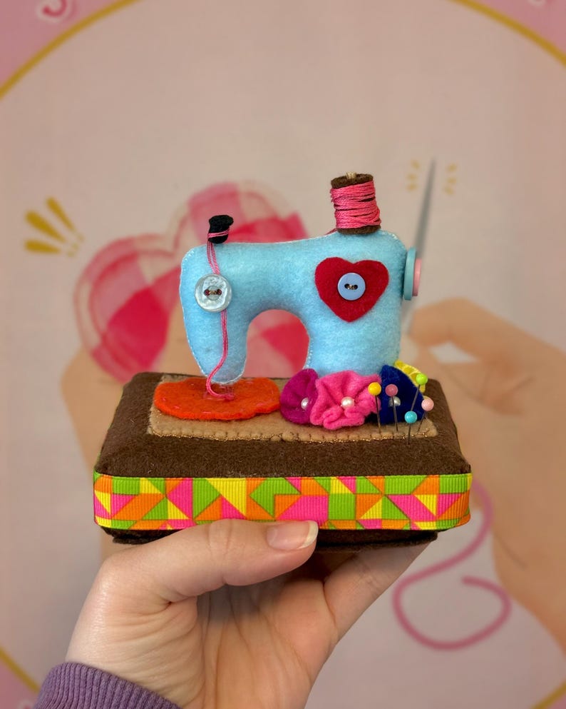 Op de afbeelding: Een handgemaakte vilten naaimachine in lichtblauw, met een rood hartdetail en een spoel roze garen. Het staat op een bruine basis met een kleurrijke geometrische lintafwerking en een klein speldenkussen met bloemspelden.