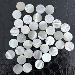May include: A collection of round, iridescent white mother-of-pearl shell discs. The discs are arranged closely together, displaying a range of natural patterns and textures. The overall effect is a shimmering, pearlescent display.