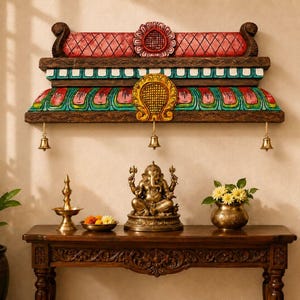 Op de afbeelding: Een houten wandplank met een decoratieve, veelkleurig gesneden kroonlijst met rode, groene en gele accenten, en drie kleine koperen belletjes aan de onderrand, geplaatst boven een donkerhouten consoletafel met een Ganesha-beeld.