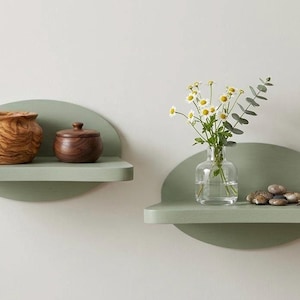 May include: Two sage green, semi-circular wall shelves. One shelf holds a wooden pot and a small wooden jar. The other shelf displays a glass vase with flowers and a small pile of stones.