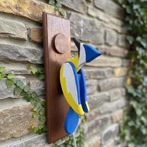 May include: A wooden blue jay wall art piece. The bird is painted with blue, yellow, and white accents and is mounted on a rectangular wooden plaque. The plaque has a circular wooden accent. The art is mounted on a stone wall.