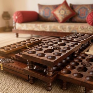 Puede incluir: Una colección de tableros de juego de madera hechos a mano, con numerosas hendiduras circulares. Los tableros están dispuestos en una exhibición en capas, mostrando intrincadas tallas y un rico acabado de madera oscura. La escena sugiere un enfoque en los juegos tradicionales y la artesanía.
