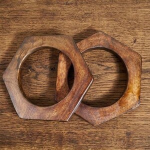 May include: Two wooden, hexagonal bangles with a natural wood grain finish. The bangles are a warm brown color and are resting on a wooden surface. The bangles are a unique accessory.